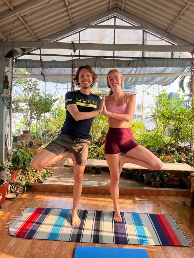 Two guests in partner tree pose on a striped rug in the garden shala