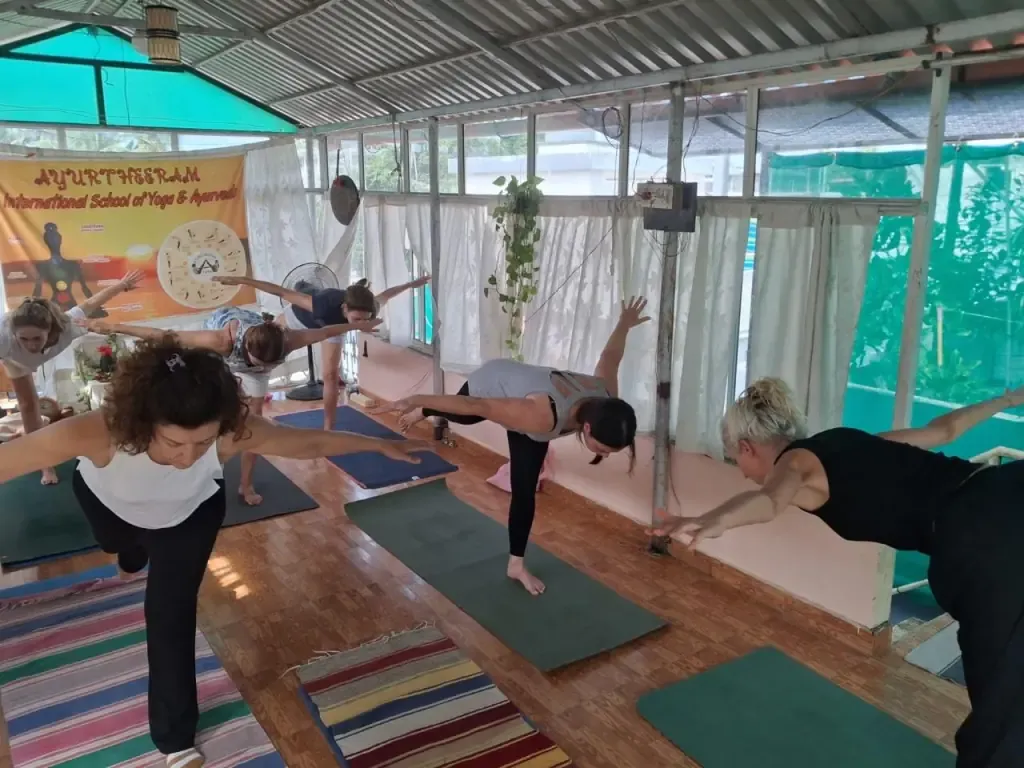 A yoga class balancing in warrior three pose on mats in the shala