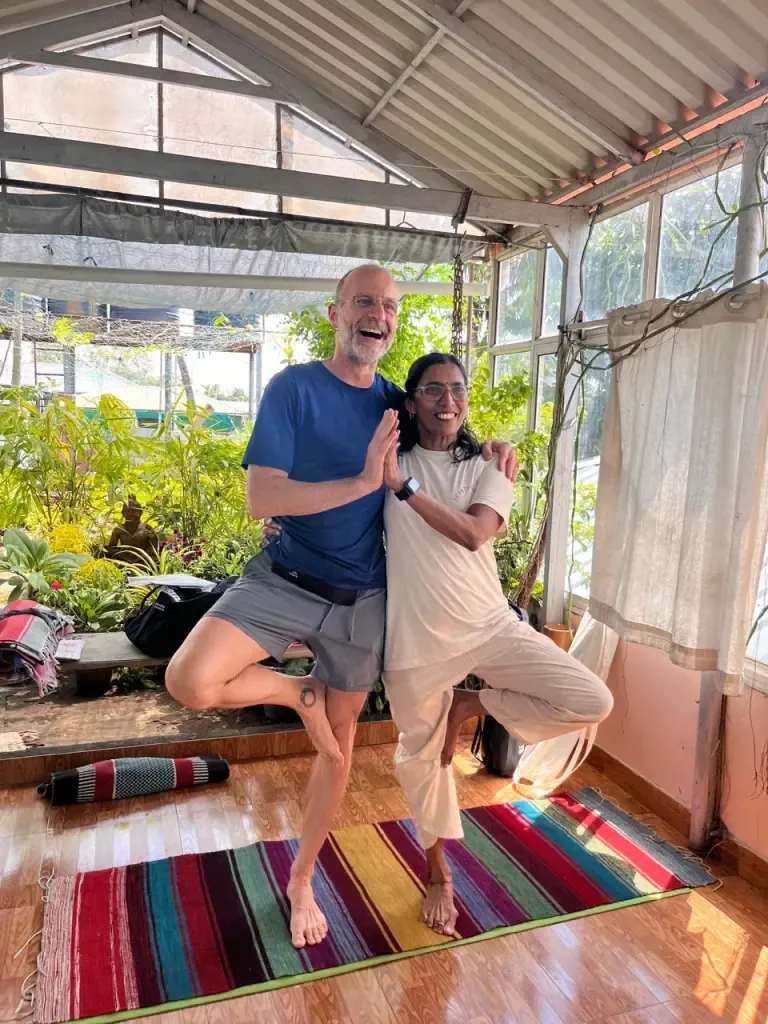 A laughing guest and a teacher in partner tree pose on a striped rug