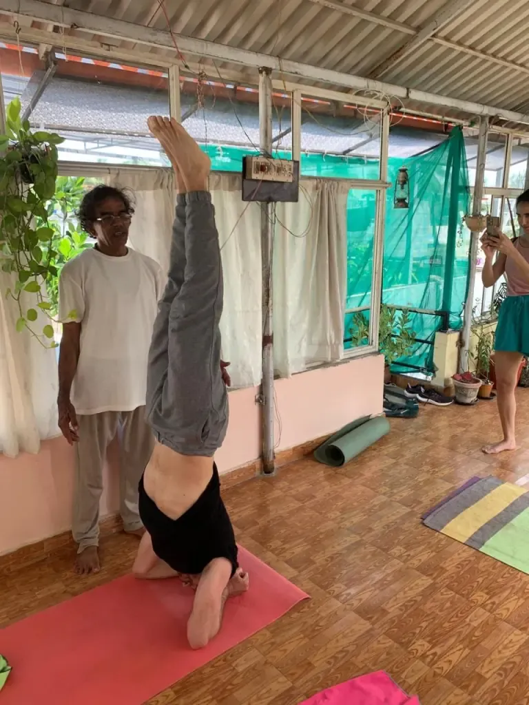 A teacher guiding a guest into a headstand by the shala windows