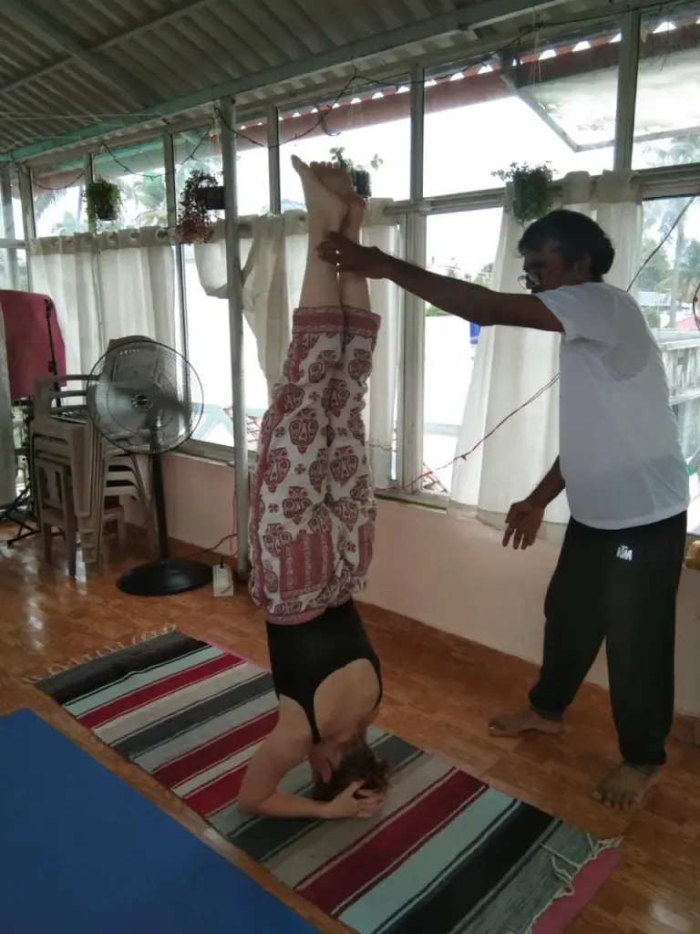 A teacher steadying a guest in a headstand beside the shala windows