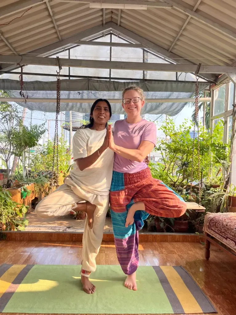 A teacher and a guest smiling in partner tree pose in the garden shala