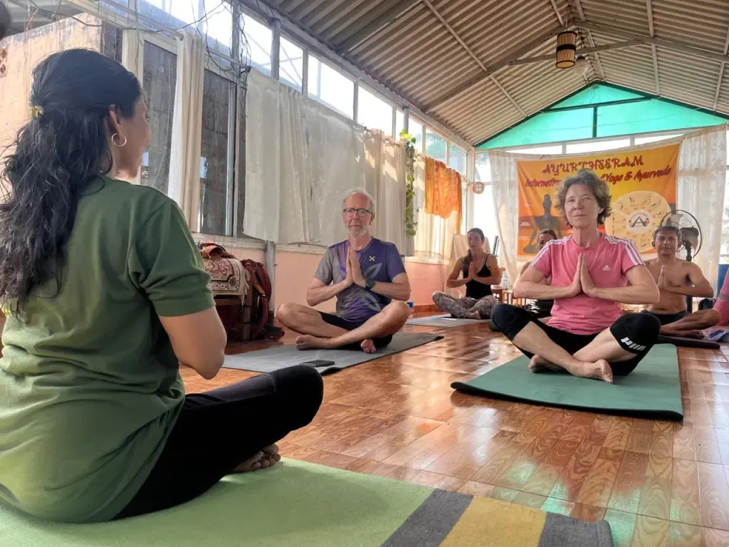 A teacher leading a seated class as guests hold their hands in namaste