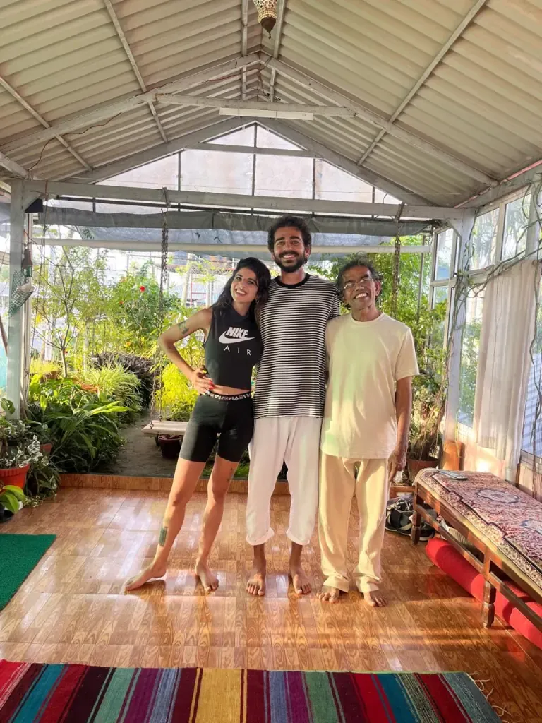 Two guests standing with their teacher in the sunny garden shala